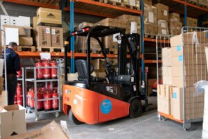In een drukke magazijnscène hint een kleine oranje vorkheftruck naar de mogelijkheden met een vorkheftruckcertificaat. Hij staat geparkeerd bij planken vol dozen, terwijl een arbeider in een blauw jasje efficiënt beweegt. Rode brandblussers staan bij elkaar op de voorgrond, terwijl er rechts nog meer dozen netjes zijn opgestapeld.