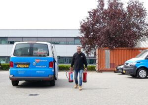 Een persoon die over een parkeerplaats loopt, met twee brandblussers in zijn handen, is een voorbeeld van waarom iedereen een BHV-cursus zou moeten volgen. Een blauwe bestelwagen met het opschrift INSTRUCTEURS staat vlakbij geparkeerd, naast een ander voertuig en een zeecontainer, met een boom en gebouw als achtergrond.