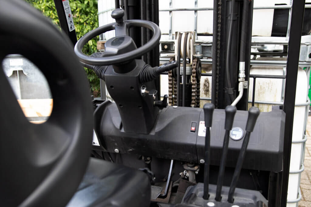 Close-up van het bedieningsgedeelte van een vorkheftruck, met de nadruk op het stuur, de hendels en de versnellingen. Op de industriële achtergrond zijn zichtbare containers te zien. Inschrijven voor een heftruckcursus? Dit zijn de opties die je nodig hebt om je vaardigheden in deze dynamische omgeving te verbeteren.