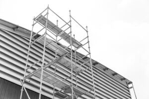 Een monochrome afbeelding legt een steigerconstructie vast die tegen de zijkant van een gebouw met horizontale gevelbekleding is geplaatst, en vraagt zich af of je op een rolsteiger zonder certificaat kunt werken. De bewolkte lucht doemt op op de achtergrond.