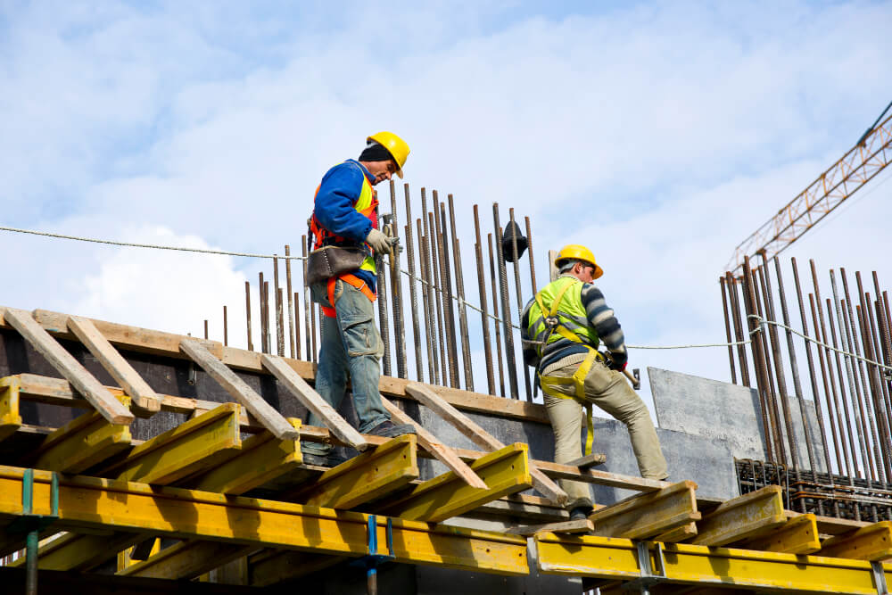 Twee bouwvakkers, uitgerust met helmen en veiligheidsmiddelen, laten de praktische voordelen van een VCA-cursus zien terwijl ze vakkundig manoeuvreren op een houten platform, omgeven door metalen staven en balken, onder een gedeeltelijk bewolkte hemel.