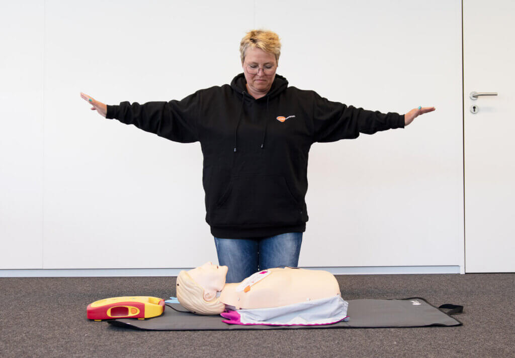 Een persoon in een zwarte hoodie staat met uitgestrekte armen en demonstreert een procedure met een reanimatiepop op de grond. Een defibrillator is naast de pop geplaatst - een praktische reden om te overwegen Redenen om een BHV-opleiding te volgen. De scène speelt zich af tegen een effen witte muur.