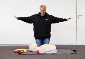Een persoon in een zwarte hoodie staat met uitgestrekte armen en demonstreert een procedure met een reanimatiepop op de grond. Een defibrillator is naast de pop geplaatst - een praktische reden om te overwegen Redenen om een BHV-opleiding te volgen. De scène speelt zich af tegen een effen witte muur.