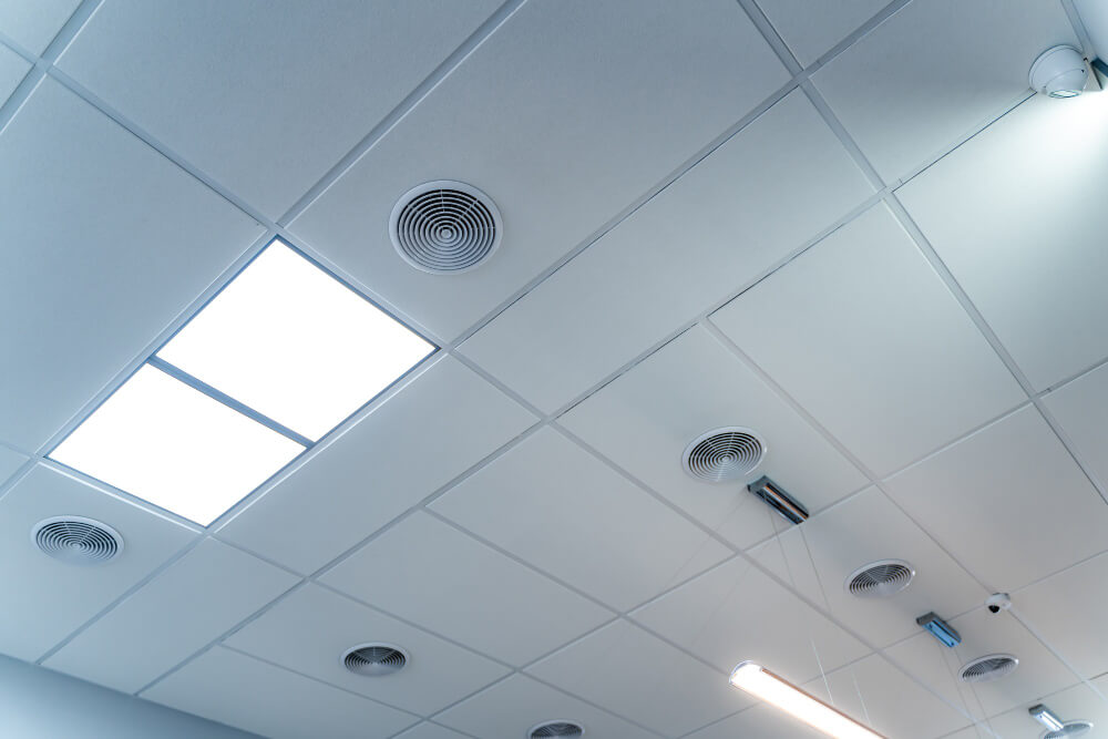 Plafond met witte panelen, verzonken ronde ventilatieopeningen en rechthoekige ledlampen tonen een modern design. Sommige plafondspots zijn ook zichtbaar: een ideale setting om ervoor te zorgen dat uw brandmeldinstallatie goed is geregeld voor optimale veiligheid en functionaliteit.