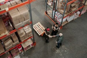 Vanuit de lucht zijn drie mensen te zien in een magazijn. Twee schudden elkaar de hand bij planken vol dozen en rode emmers, misschien om een deal te sluiten over certificeringsvereisten. Een palletwagen staat in de buurt, wat duidt op discussies over EPT-behoeften en -procedures.