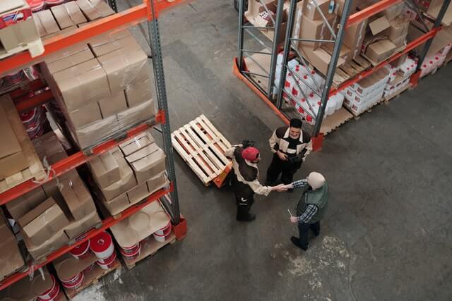 Vanuit de lucht zijn drie mensen te zien in een magazijn. Twee schudden elkaar de hand bij planken vol dozen en rode emmers, misschien om een deal te sluiten over certificeringsvereisten. Een palletwagen staat in de buurt, wat duidt op discussies over EPT-behoeften en -procedures.