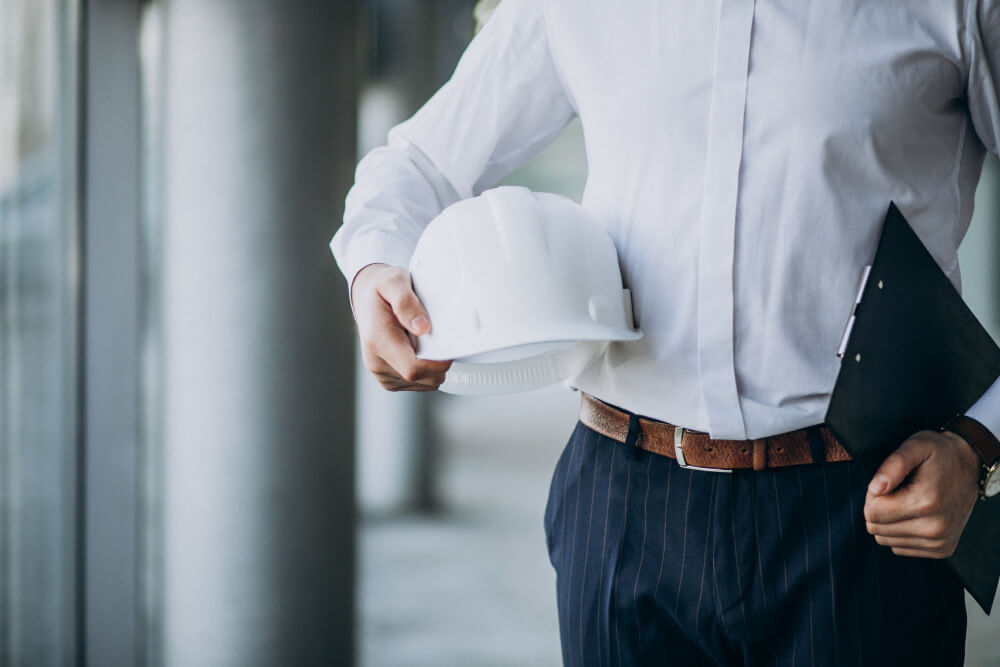 Een persoon in een wit overhemd en een gestreepte broek houdt een witte helm in de ene hand en een zwart klembord in de andere, staande bij grote ramen. Je ziet een vacature preventiemedewerker: wat houdt deze functie in? Het suggereert een rol gericht op veiligheid en organisatie binnen een professionele setting.
