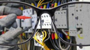 Close-up van een bril die focust op een elektrisch paneel met draden en schakelaars. Ter ere van De internationale dag van het zicht, is een persoon met handschoenen bezig met het aanpassen van een onderdeel met een schroevendraaier. Het montuur van de bril is prominent op de voorgrond, waardoor het zicht gedeeltelijk wordt belemmerd.