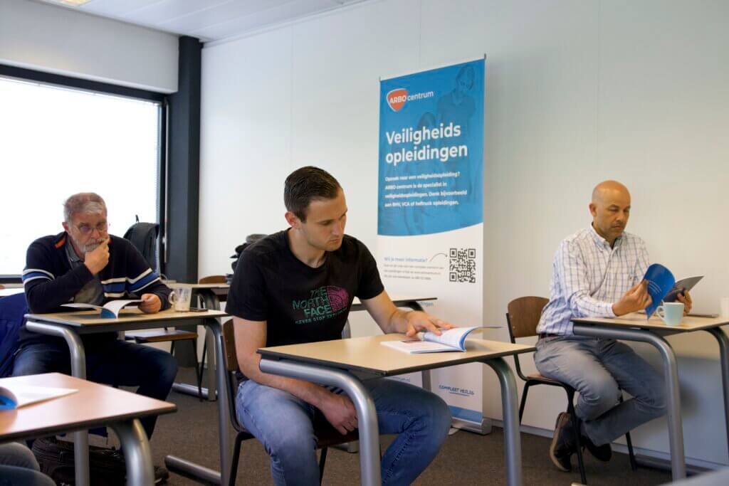 Drie mannen zitten aan bureaus in een klaslokaal, verzonken in boekjes. Een banner op de achtergrond benadrukt veiligheidstraining, en benadrukt waarom bedrijfscultuur van invloed is op de veiligheid op de werkplek. Eén man draagt een zwart T-shirt, een ander een geruit overhemd en de derde een blauwe trui.