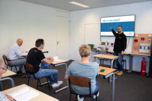 Een leraar staat voor een klaslokaal en wijst naar een scherm met de tekst: Het belang van regelmatige veiligheidstrainingen voor werknemers. Vier zittende studenten luisteren aandachtig, omringd door tafels, stoelen en leermiddelen.