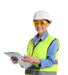 Met een witte helm, een gele veiligheidsbril en een neongeel vest over een blauw shirt is deze persoon een voorbeeld van het belang van het gebruik van persoonlijke beschermingsmiddelen op het werk. Glimlachend houden ze een klembord en een pen vast en inspecteren of maken ijverig aantekeningen.