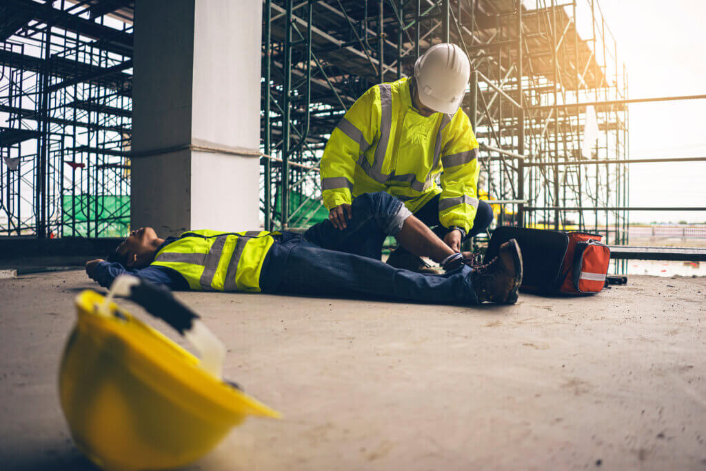 Een bouwvakker verzorgt een collega die op de grond ligt, mogelijk gewond, op een bouwplaats met steigers. De staande werknemer, die laat zien hoe je onverwachte noodsituaties op de werkplek aanpakt, draagt een geel veiligheidsvest en een helm. Een gele helm en EHBO-doos liggen in de buurt.