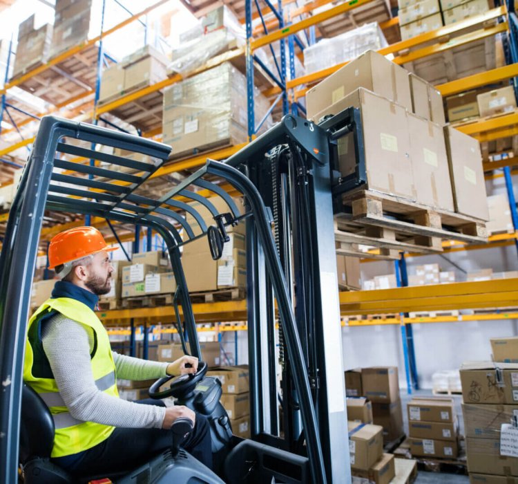 Een werknemer in een fluorescerend vest en een oranje helm bedient een vorkheftruck en tilt een pallet dozen op in een groot magazijn. In Nederland staan planken vol met gestapelde dozen op de achtergrond, wat laat zien hoe heel Nederland efficiënt werkt om de toeleveringsketen draaiende te houden.