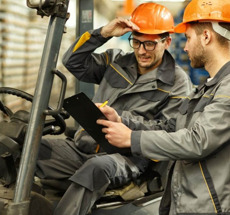 Twee bouwvakkers in grijze uniformen en oranje helmen bespreken een test aanvraag terwijl ze een klembord vasthouden. De een zit op een heftruck, de ander staat naast hem in een industriële setting met planken op de achtergrond.
